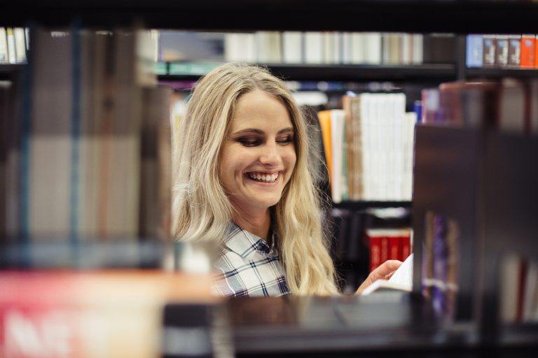 Jeune femme souriante lisant un livre dans une bibliothèque, entourée d'étagères remplies de livres. | Transit-Jeunesse - Programme bourse